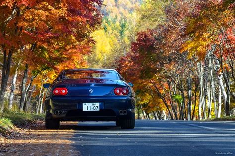 a blue car driving down a road surrounded by trees with fall foliage on ...