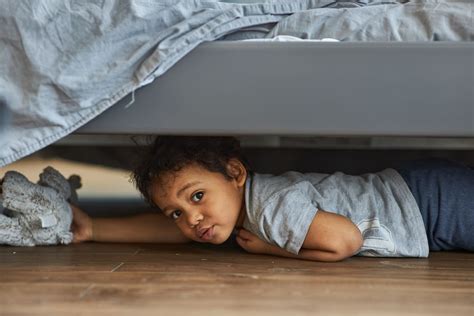 Family at home concept, boy playing hide and seek. Cute afro boy peeking out from under the bed ...