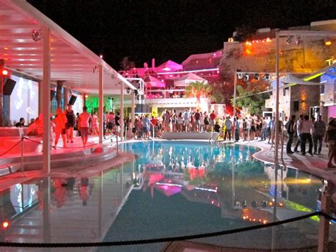 a group of people standing around in front of a pool at night with lights on