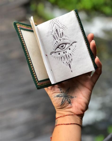 a hand holding an open book with the eye drawn on it's cover, in front of a river