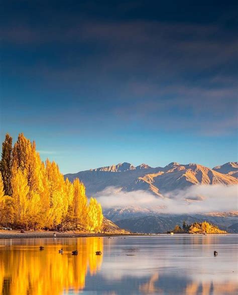 a lake surrounded by mountains with trees in the foreground and clouds ...