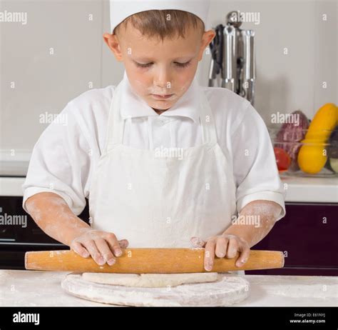 Young boy in chefs uniform rolling dough with a large wooden rolling pin as he prepares the base ...