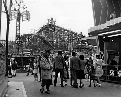 black and white photograph of people standing in front of roller coaster at an amusement park