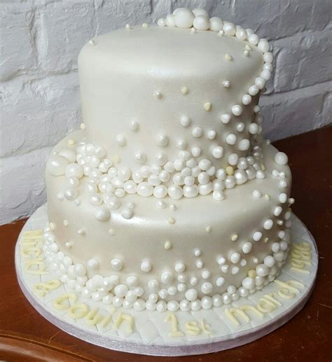 a three tiered white cake with pearls on it sitting on a table next to a brick wall