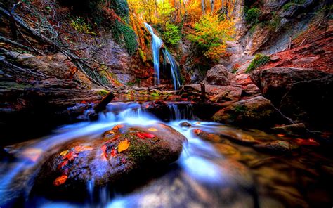 a small waterfall in the middle of a forest filled with trees and rocks, surrounded by fall foliage