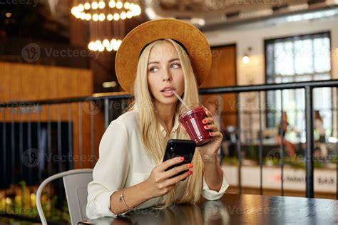Pensive blue eyed young blonde trendy woman in stylish clothes sitting over modern cafe interior ...