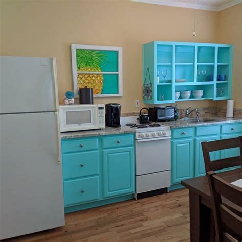 a kitchen with blue cabinets, white appliances and a pineapple painting on the wall