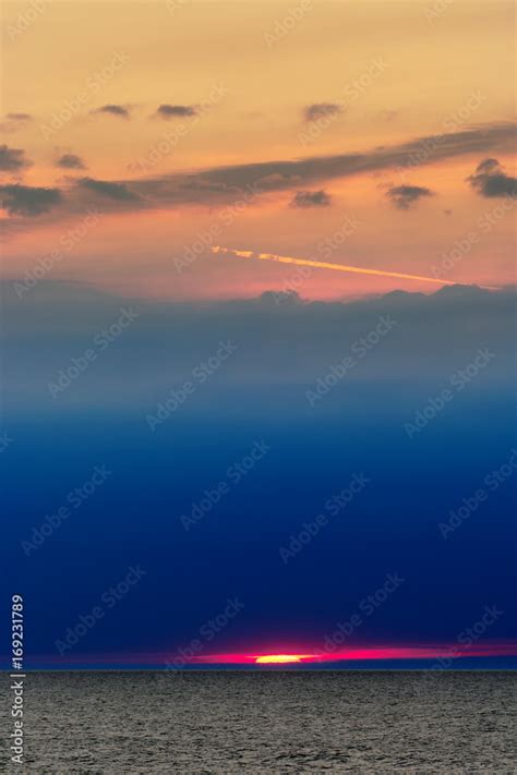 Unusual sunset at sea with sun partly seen through thick layer of dark clouds. Seascape at dusk ...