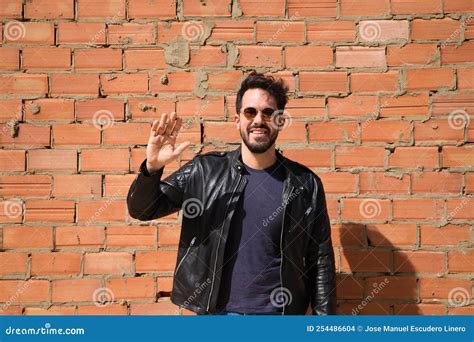 Young Latino Man, Dark with Beard and Sculpted Body Greets with Raised Hand. Red Brick Wall in ...