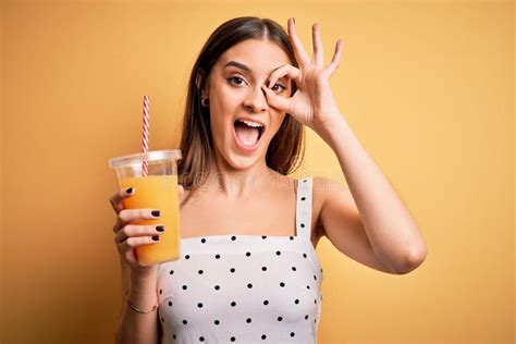 Young Beautiful Brunette Woman Drinking Healthy Orange Juice Over Yellow Background with Happy ...