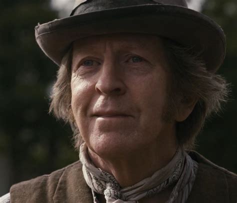 an older man wearing a brown vest and hat looking off to the side with trees in the background