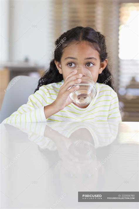 Little girl drinking water from glass at table at home and looking away — refreshment, female ...