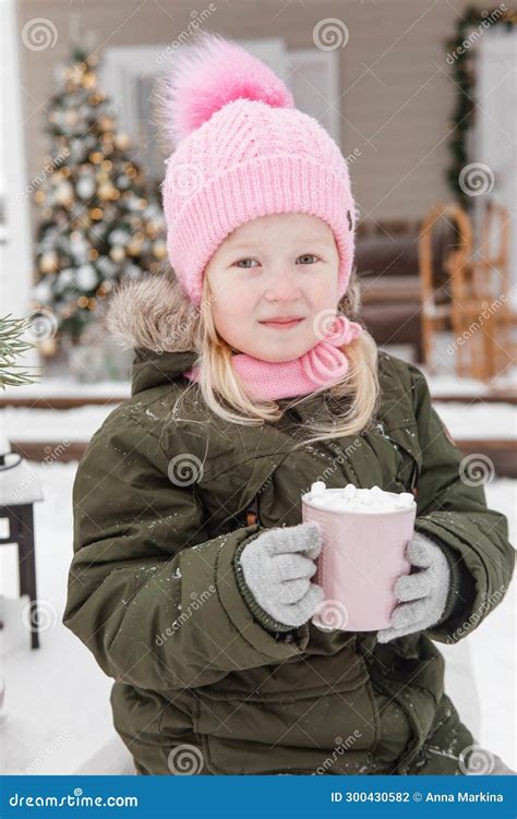 A Little Girl in Winter Clothes with a Cup of Hot Cocoa with Marshmallows, in the Courtyard of a ...