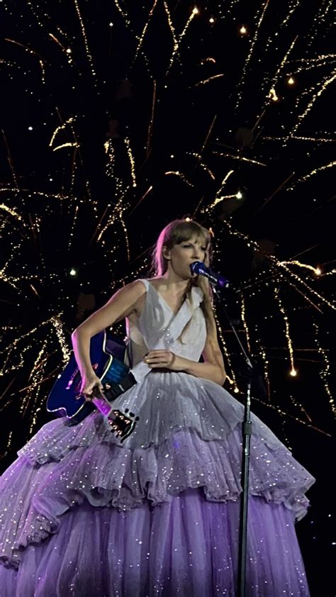 a woman in a purple dress holding a guitar and singing into a microphone with fireworks behind her