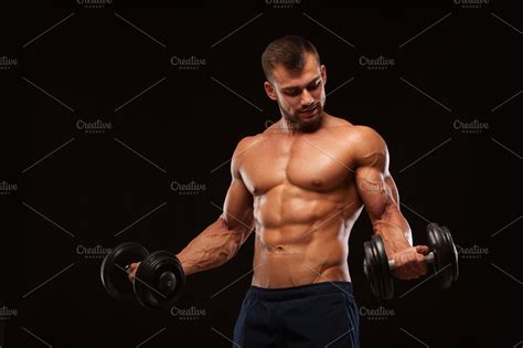 Handsome athletic man in gym is pumping up muscles with dumbbells in a ...