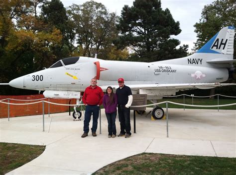 two men and a woman standing in front of a navy jet plane at an air museum