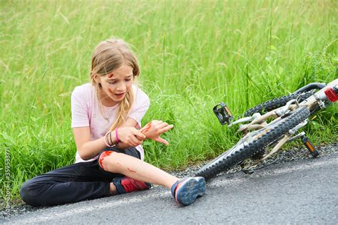 Sad crying little child girl fell from the bike in the summer park. Bleeding on hands and feet ...