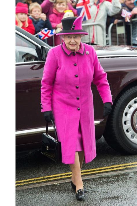 an older woman in a pink coat and hat walks past a car as people watch