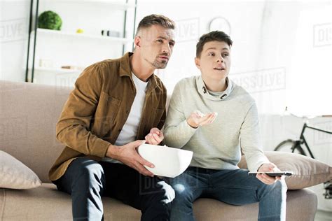 Serious Father And Teen Son Watching Tv And Holding Bowl Of Popcorn At