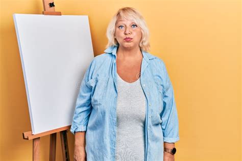 Middle Age Blonde Woman Standing By Painter Easel Stand Puffing Cheeks