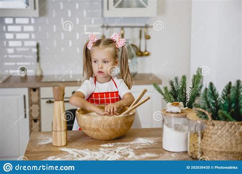 Four Year Old Blonde Girl In A White T Shirt And Red Apron Kneads Dough At Home In The Kitchen