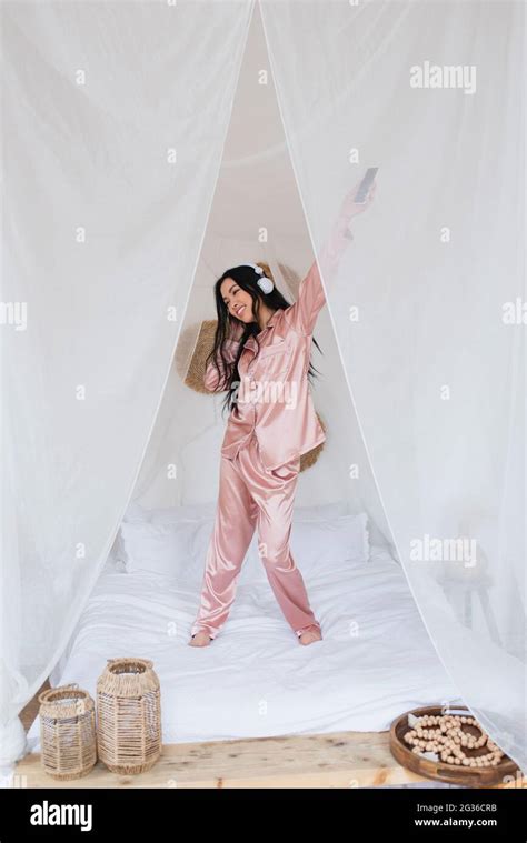 Positive Young Asian Woman Dancing On Bed With Headphones And Holding