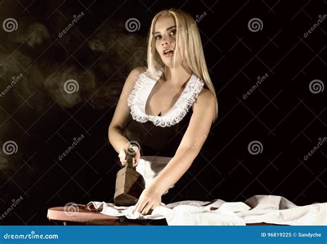 Blonde Woman In Housekeeper Suit Ironing White Shirt With Old Iron Retro Style On A Dark