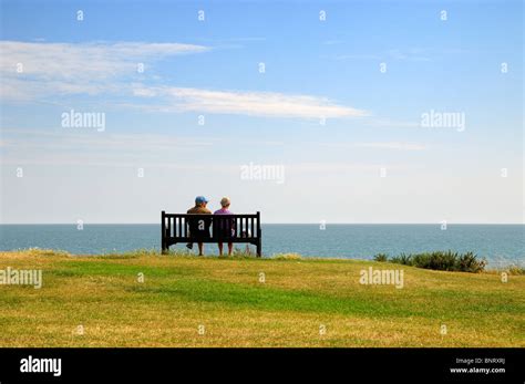 Back View Of Elderly Couple Sitting On Cliff Bench Looking Out To Sea