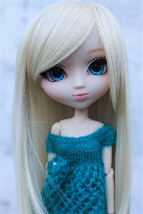 A Doll With Blonde Hair And Blue Eyes Wearing A Knitted Dress In Front Of A White Wall