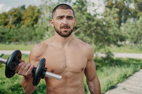 Outdoor Shot Of Muscular Bodybuilder With Naked Torso Raises Barbell And Has Strong Biceps