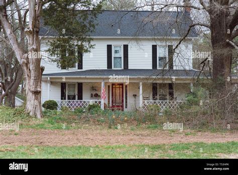 A charming white country farmhouse with black shutters sits in the woods in a rural setting ...