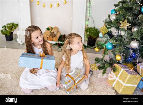 Zwei kleine blonde Mädchen in weißen Kleidern spielen am Kamin und am Weihnachtsbaum mit Kisten