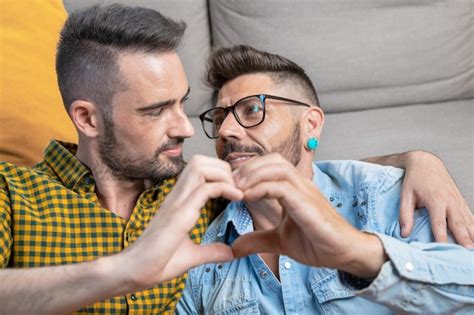 Joven pareja gay sonriendo confiada haciendo el símbolo del corazón con las manos en casa Foto