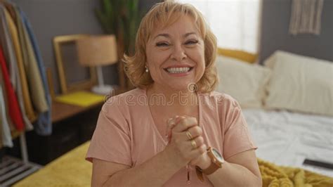 A Beautiful Mature Blonde Caucasian Woman Smiles Joyfully While Seated In Her Cozy Bedroom