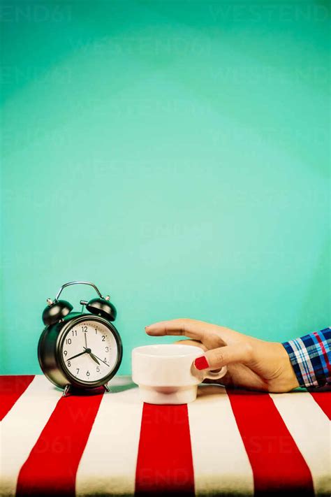 Hand Of Woman Holding Cup In Front Of Old Fashioned Alarm Clock Stock Photo