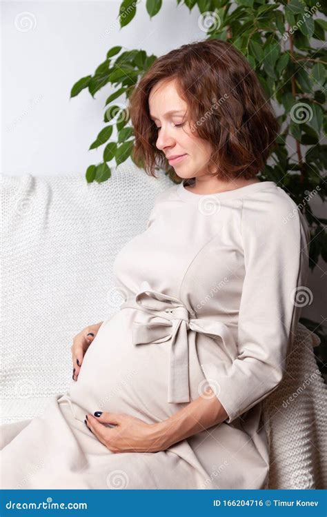 Portrait Of Young Beautiful Curly Pregnant Woman In Beige Nude Dress Holding Hands On Her Tummy