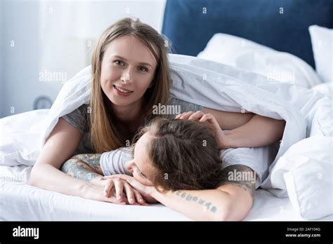 Two Smiling Lesbians Embracing While Lying Under Blanket In Bed Stock