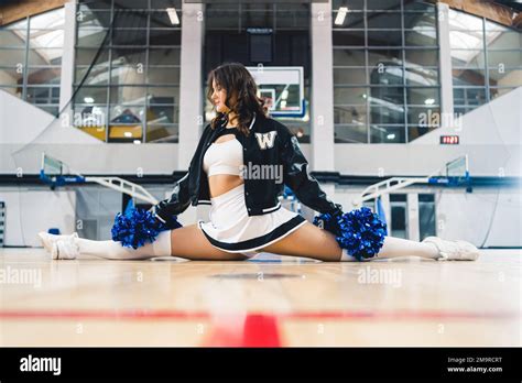 Front View Of A Brunette Cheerleader Holding Blue Shiny Pom Poms And Doing A Split On Basketball