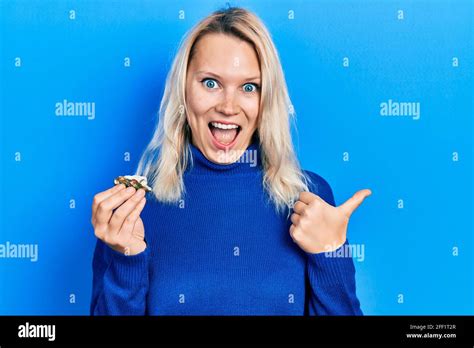 Beautiful Caucasian Blonde Woman Holding Raw Oyster Pointing Thumb Up To The Side Smiling Happy
