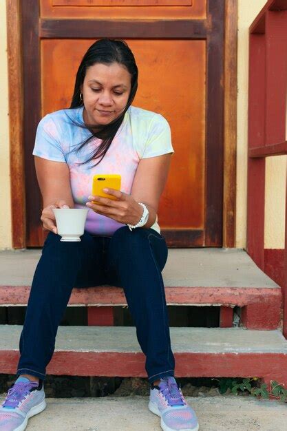 Linda latina sentada do lado de fora de casa tomando café e verificando o celular Foto Premium