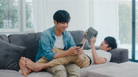 Joven pareja gay lgbtq utilizando teléfono móvil y tableta en casa moderna Hombre asiático