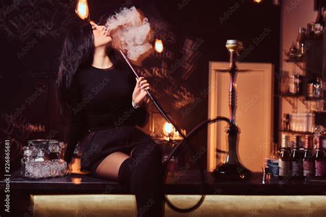 Sexy Brunette Girl In Seductive Black Clothes Smokes A Hookah While Sitting On Counter In A