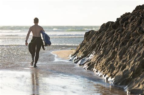 Premium Photo A Man With A Naked Torso Walking On The Beach Holding A Surfboard View From The Back
