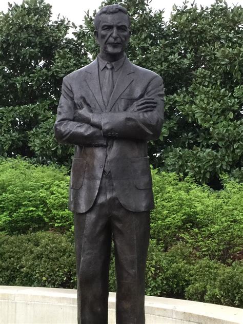 A Statue Of A Man With His Arms Crossed In Front Of Some Bushes And Trees