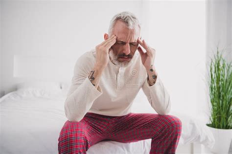 Mature Man Experiencing Morning Head Pain While Sitting On Bed In Bright Bedroom Stock Photo