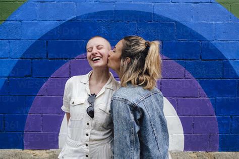 Happy Woman Kissing On Cheeks Of Non Binary Friend In Front Of Colorful