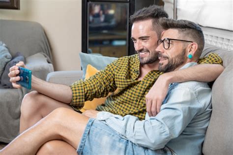 Alegre pareja gay sentada en el sofá abrazándose y mirando el teléfono móvil viendo una película