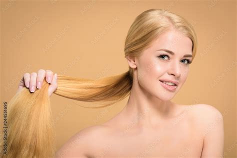 Beautiful Naked Blonde Woman Holding Long Hair And Smiling At Camera Isolated On Beige Stock