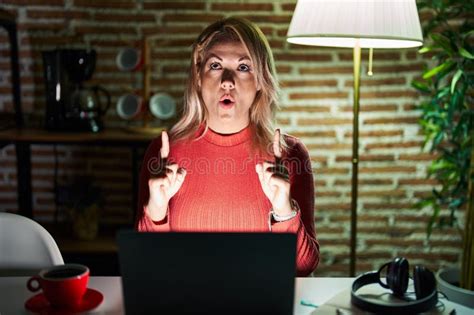 Blonde Woman Using Laptop At Night At Home Amazed And Surprised Looking Up And Pointing With