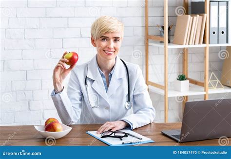 Smiling Woman In White Coat Holds Red Apple Sitting At Table In